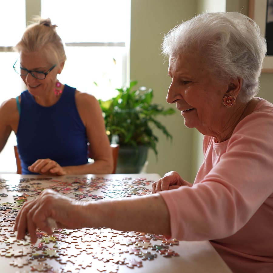 Baxter Senior Living | Happy smiling, laughing elderly senior woman sitting at table with mature middle aged daughter working on puzzle